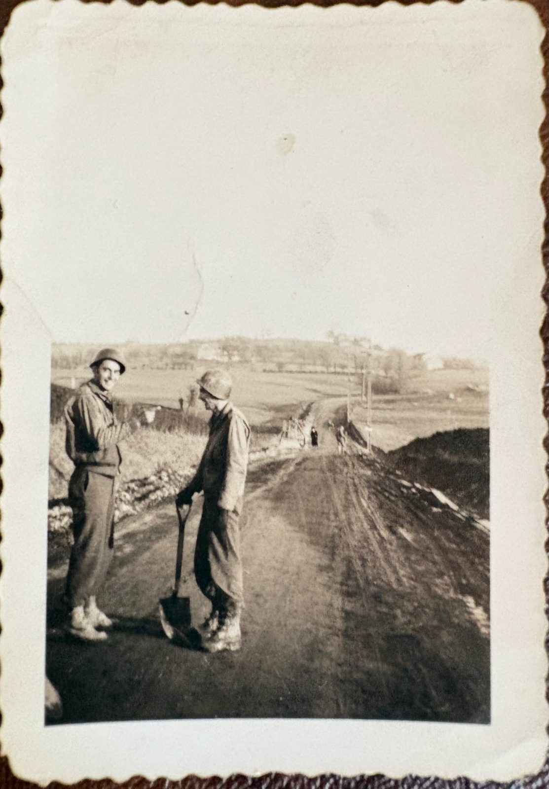 Two engineers on a dirt road, one with a shovel, a hilltop village beyond — road work in the European countryside.